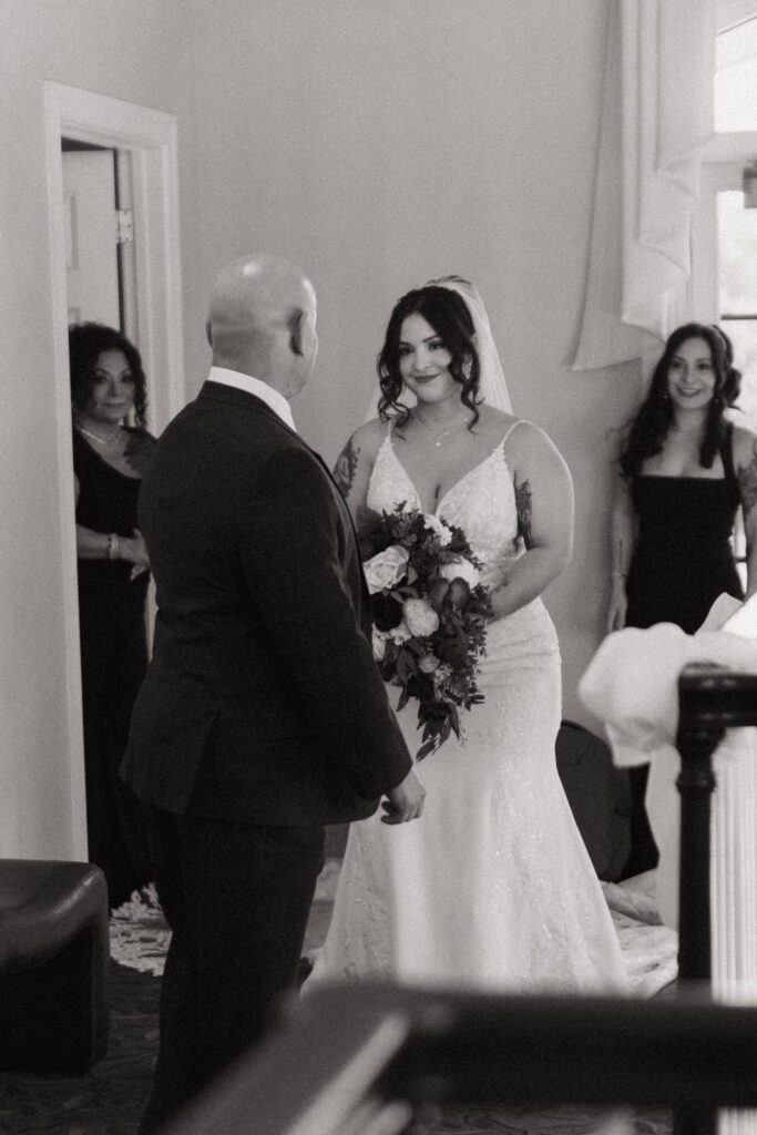 A bride having a first look with her father. While her mother and sister smile at Black Swan Country Club in Georgetown, Massachusetts.