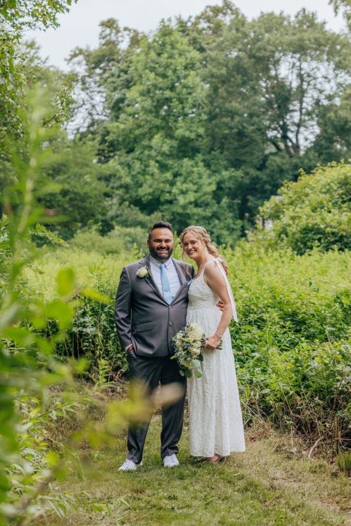A bride and groom smiling in whimsical tall grass in the woods at Bird Park in Walpole, Massachusetts during their summer elopement.