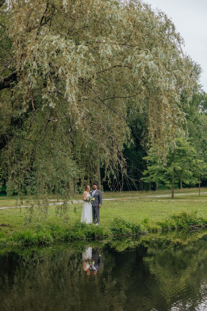 Bride and groom standing under willow tree smiling in front of pond at Bird Park in Walpole, Massachusetts.