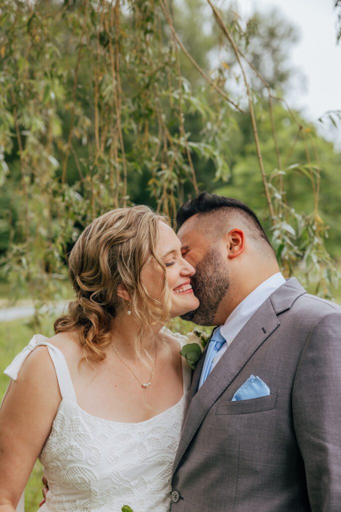 Groom kissing bride on the cheek under willow tree at Bird Park in Walpole, Massachusetts.