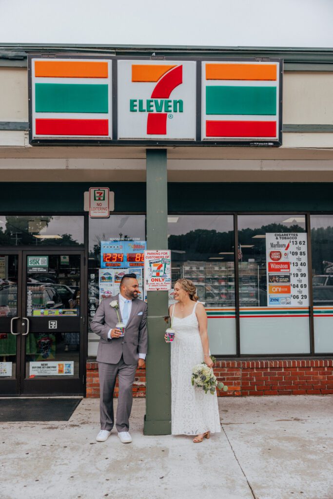 Bride and groom smiling at each other as they lean against pole at 7/11 on their wedding day.