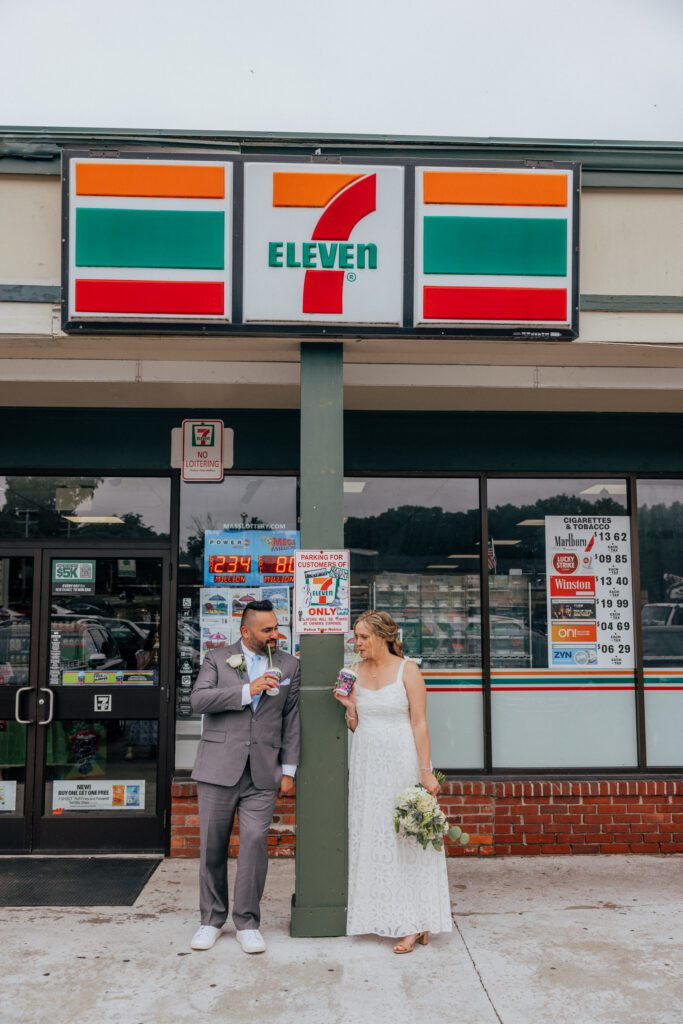 Bride and groom drinking slushies and looking at each other as they lean against pole at 7/11 on their wedding day.