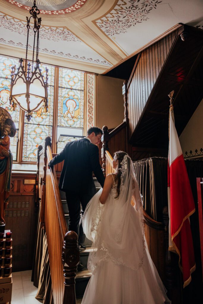 Bride and groom walking up stair case in front of stained glass at Quinet Saint John Garden of Our Lady in Massachusetts.