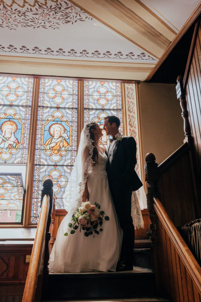 Bride and groom smiling at each other on staircase in front of stained glass at Quinet Saint John Garden of Our Lady in Massachusetts.