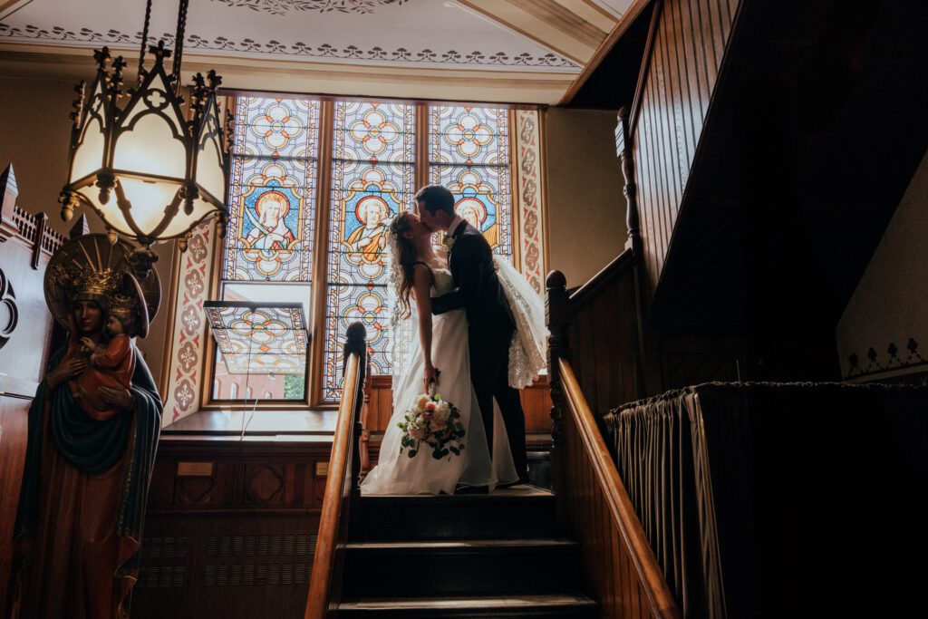 Bride and groom romantically kissing on stair case in front of stained glass at Quinet Saint John Garden of Our Lady in Massachusetts.