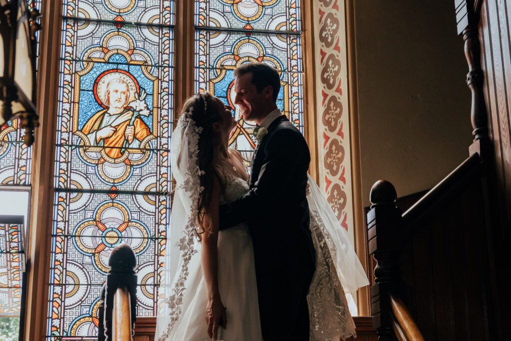 Bride and groom laughing on staircase in front of stained glass at Quinet Saint John Garden of Our Lady in Massachusetts.