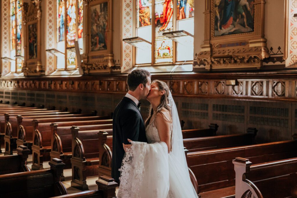 Bride and groom romantically kissing in church with stained glass at Quinet Saint John Garden of Our Lady in Massachusetts.