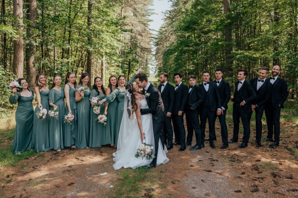 Bride and groom kissing in front of bridal party in the woods in Massachusetts.