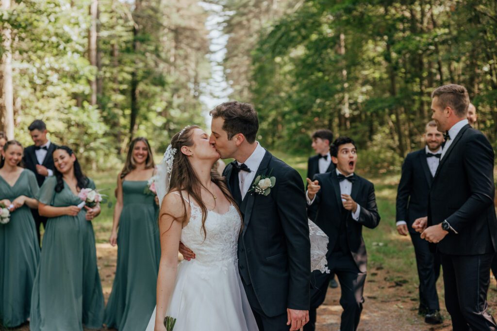 Bride and groom kissing in the woods with their bridal party cheering in the background in Massachusetts.