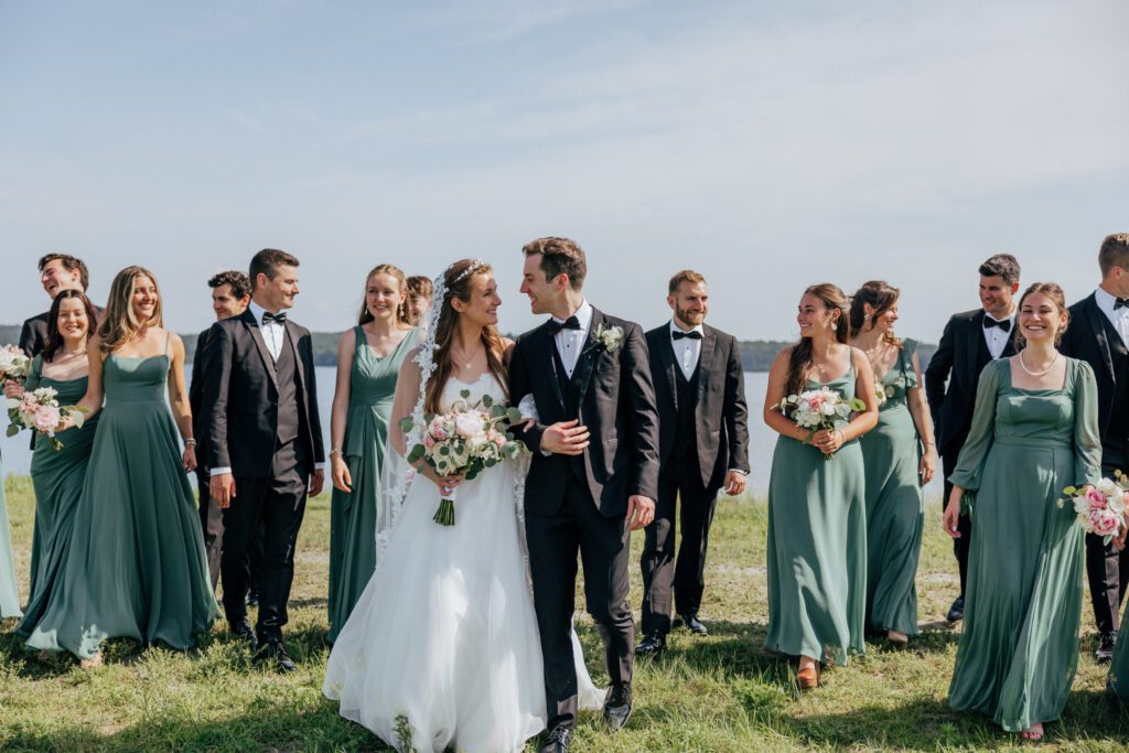 Bride and groom smiling at each other with bridal party walking behind them at water front in Massachusetts.
