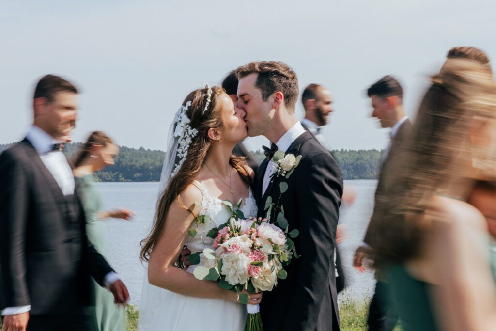 Bride and groom kissing as bridal party walks by in front of pond in Massachusetts.