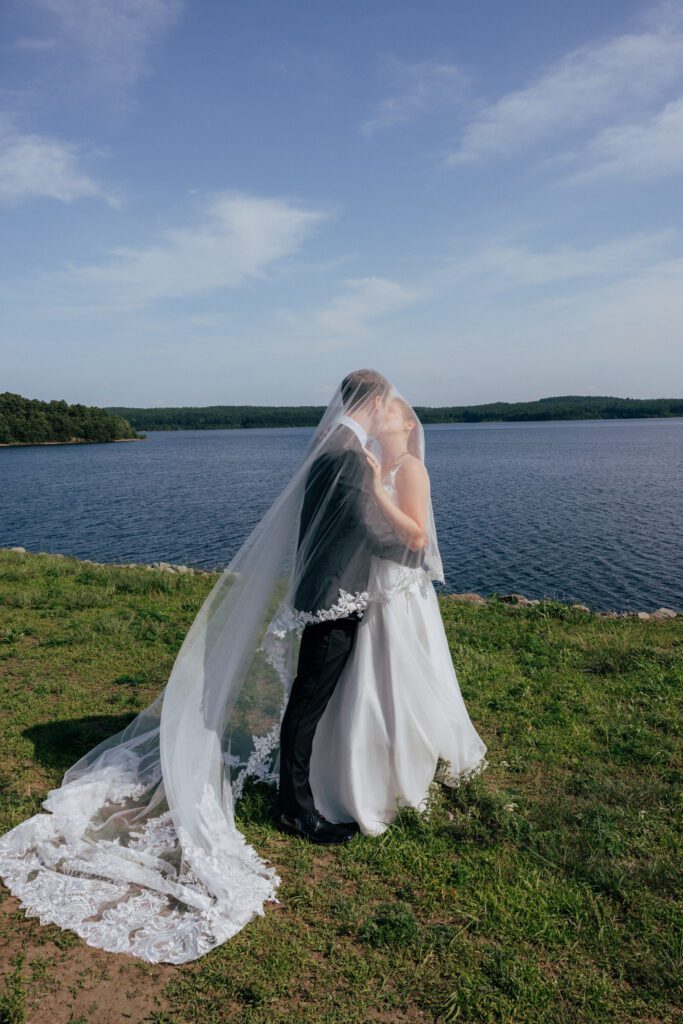 Bride and groom kissing under veil in front of waterfront in Massachusetts.