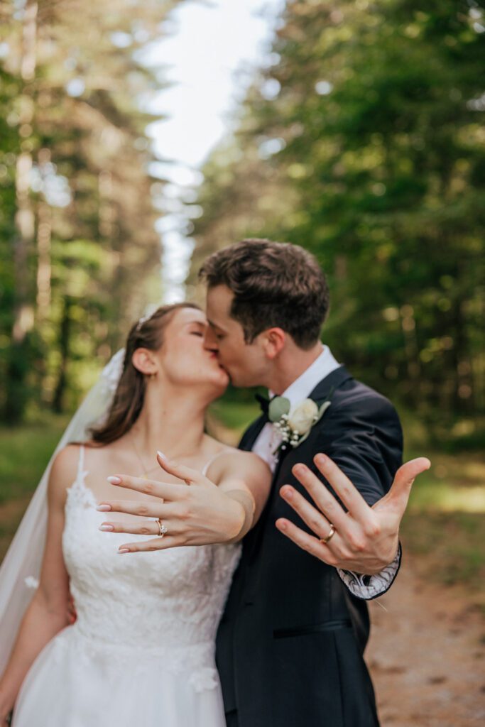 Bride and groom holding out their wedding rings while they kiss in the woods in Massachusetts.
