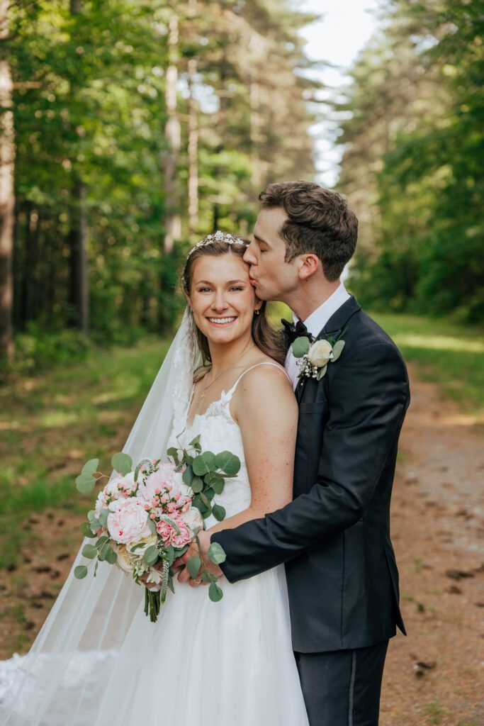 Groom romantically kisses bride on the head as she smiles in the woods in Massachusetts.