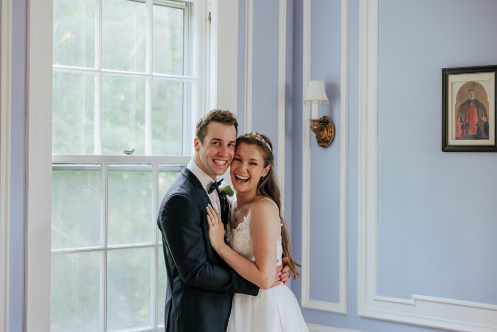 Bride and groom laughing indoors next to a window in Massachusetts.
