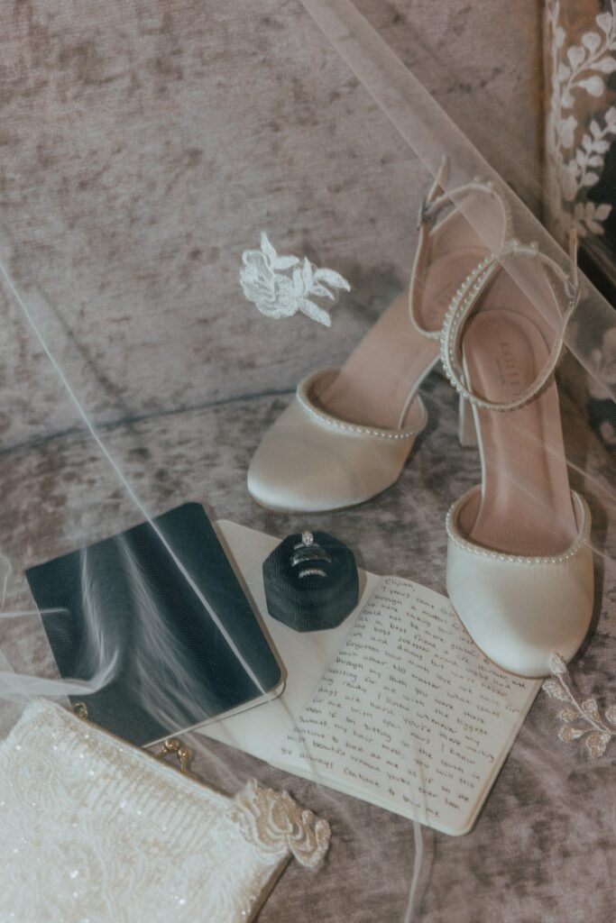 Flat lay of wedding shoes, vows, rings, and bride bag under bridal veil.