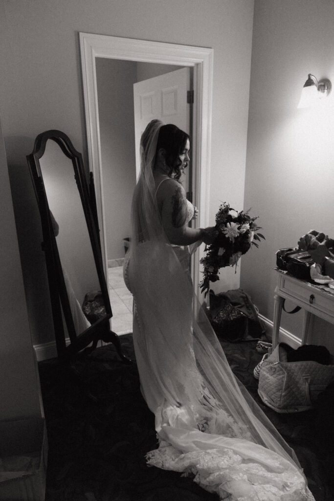 A bride getting ready in bridal suite and holding her flowers in front of mirror at Black Swan Country Club in Georgetown, Massachusetts.