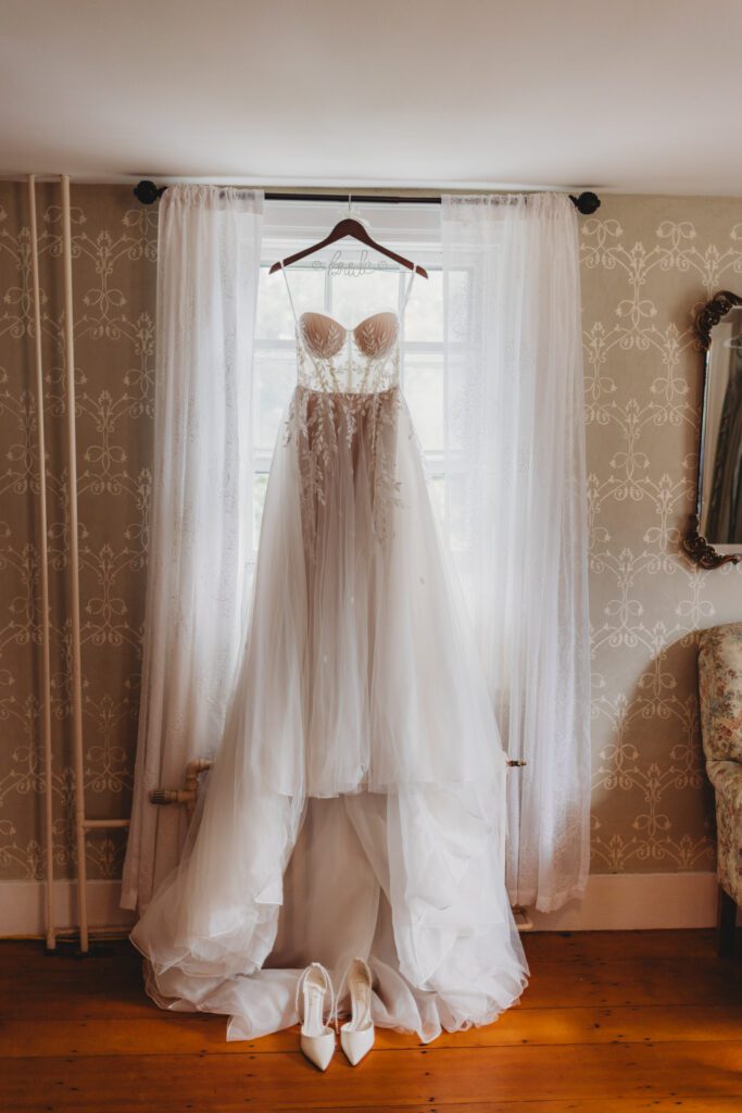 The bride's shoes and wedding dress hanging from a window in the getting ready suite at The Evermore at Peirce Farm Estate in Topsfield, Massachusetts.