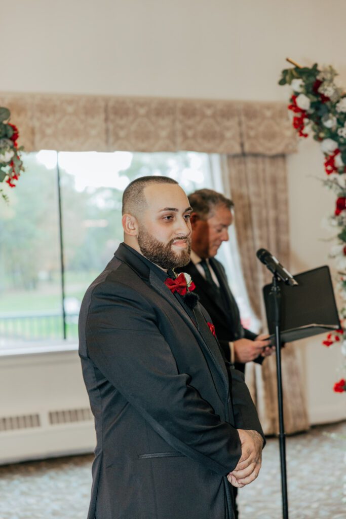 Groom smiles as bride walks down the aisle towards him at romantic indoor ceremony at Black Swan Country Club in Georgetown, Massachusetts.