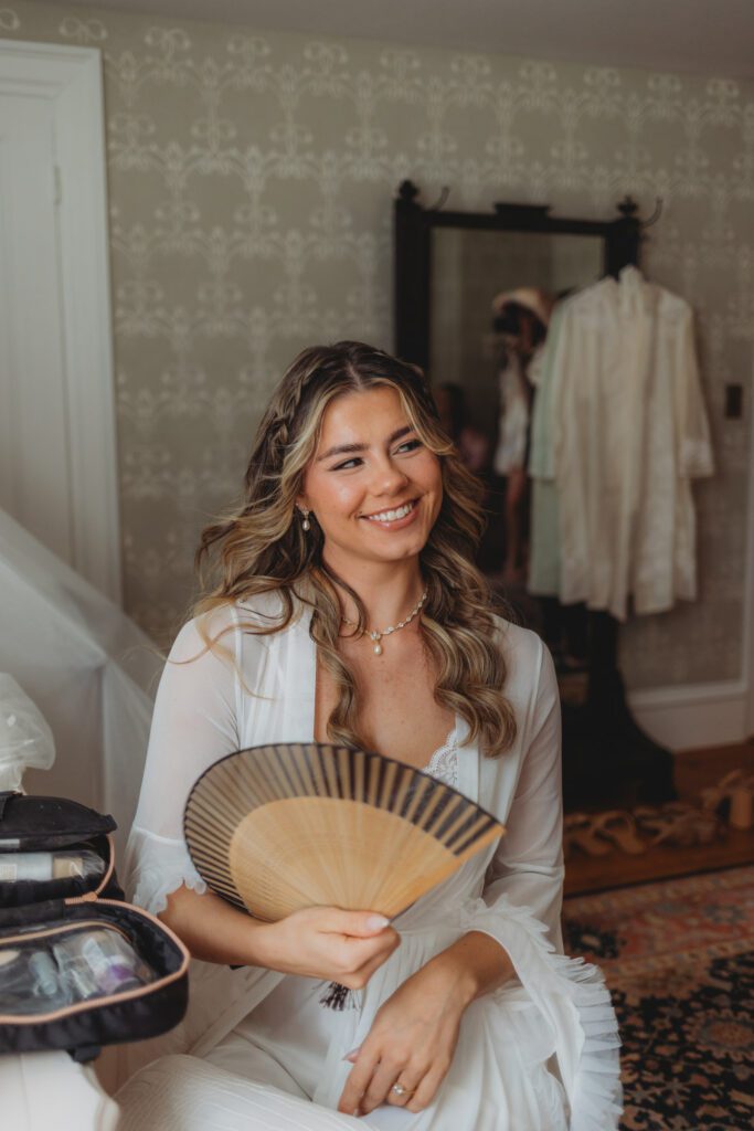 Bride smiling in a robe and fanning herself as she gets ready at The Evermore at Peirce Farm Estate in Topsfield, Massachusetts.
