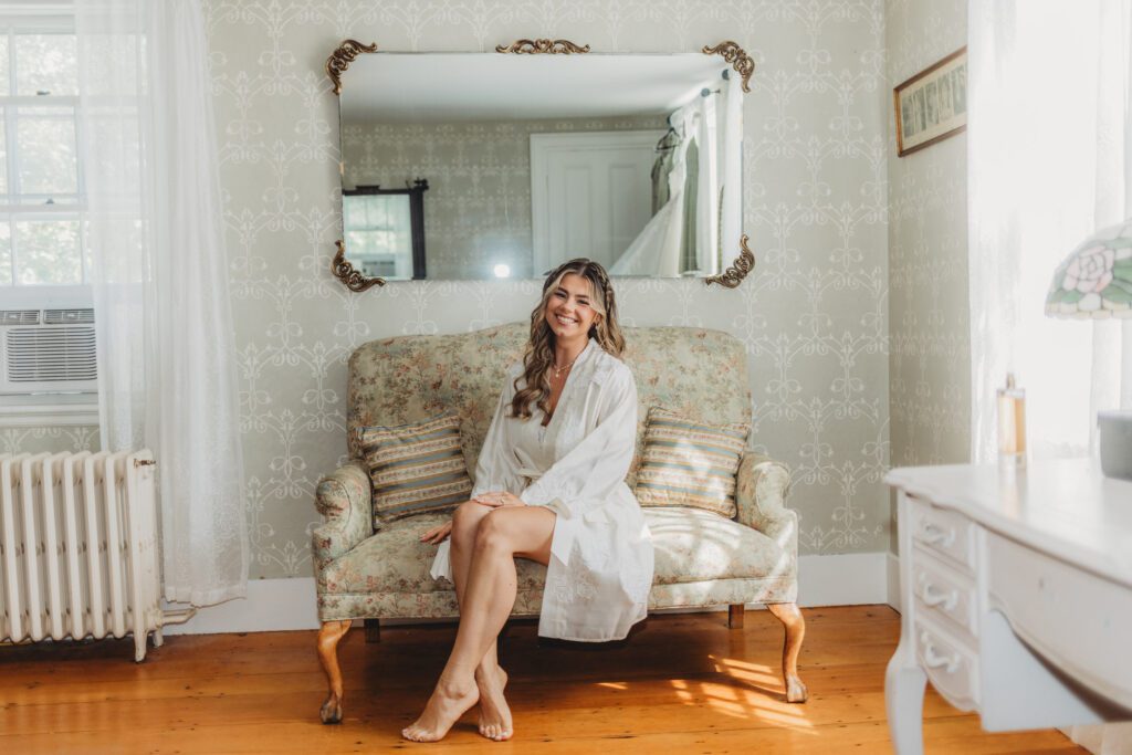 Bride sitting on a couch smiling in a silk robe in the bridal suite at The Evermore at Peirce Farm Estate in Topsfield, Massachusetts.