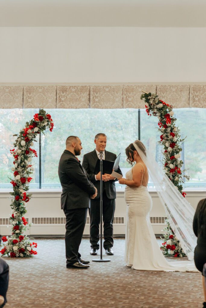 Bride an groom standing under flower arch during romantic indoor wedding ceremony at Black Swan Country Club in Georgetown, Massachusetts.