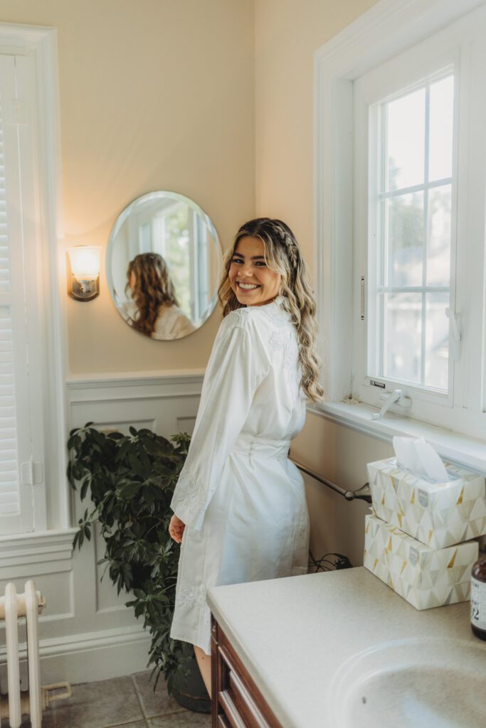 Bride in silk rob smiling over her shoulder as she gets ready at The Evermore at Peirce Farm Estate in Topsfield, Massachusetts.