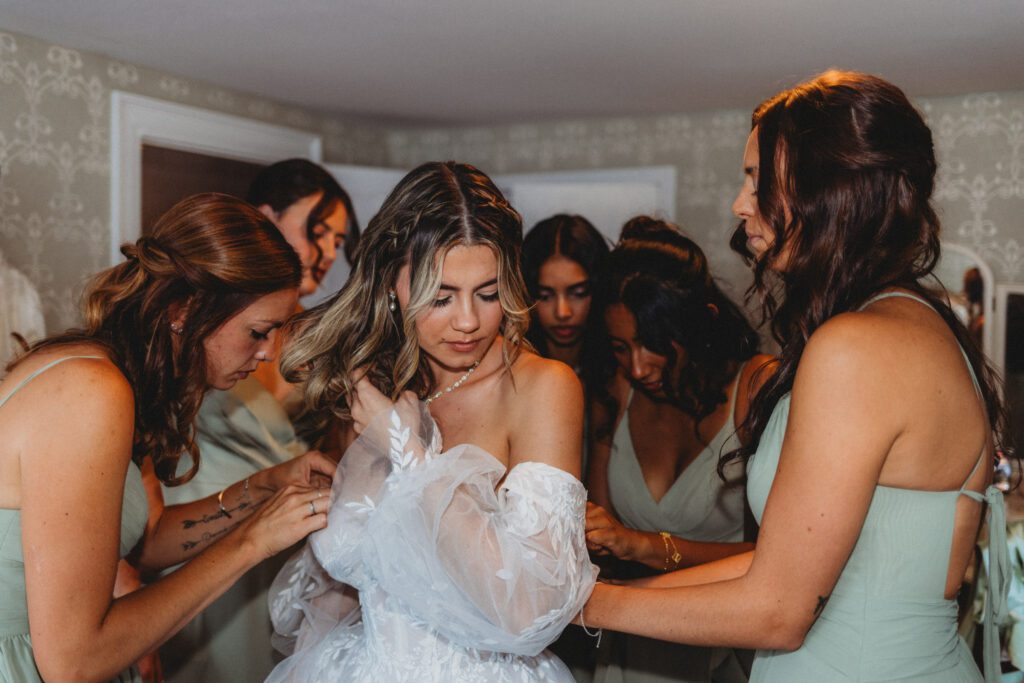 Bridesmaids helping bride put on wedding dress as she looks down at The Evermore at Peirce Farm Estate in Topsfield, Massachusetts.