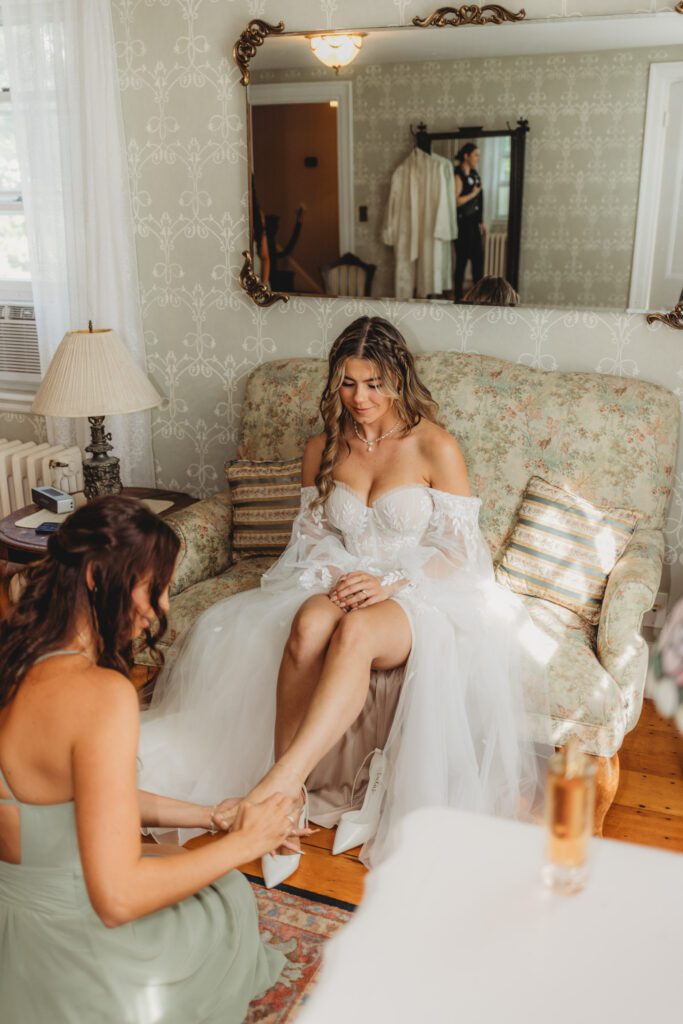 Bridesmaid putting brides shoes on as they get ready at The Evermore at Peirce Farm Estate in Topsfield, Massachusetts.