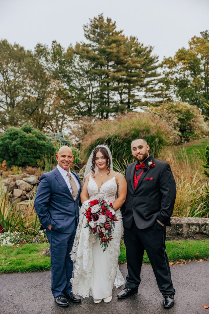 Bride, groom, and father of the bride smiling outside in front of greenery at Black Swan Country Club in Georgetown, Massachusetts.