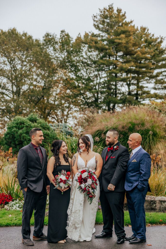 A bride and groom and family smiling at each other standing outside in front of greenery at Black Swan Country Club in Georgetown, Massachusetts.