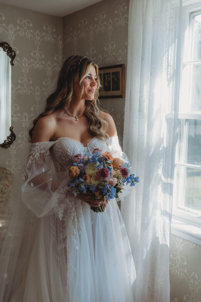 A bride looking out the window and smiling in the getting ready suite at Peirce Farm Estate in Topsfield, Massachusetts.