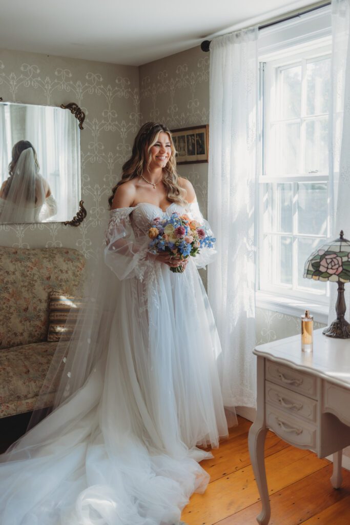 A bride laughing and holding flowers in bridal suite at Peirce Farm Estate in Topsfield, Massachusetts.
