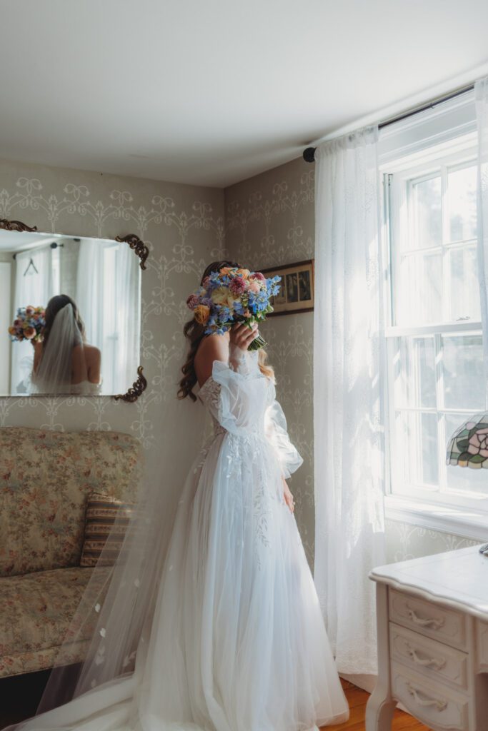 A bride covering her face with flowers in front of window at Peirce Farm Estate in Topsfield, Massachusetts.