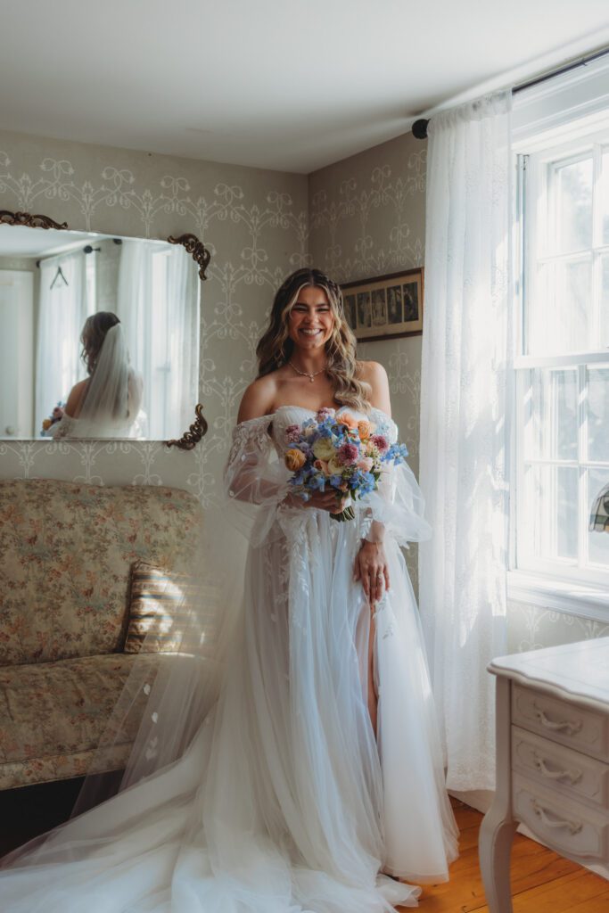 Bride smiling and holding flowers as she rests her hand on her leg in the bridal suite at Peirce Farm Estate in Topsfield, Massachusetts.