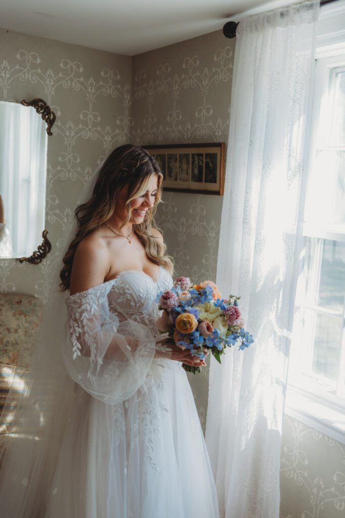 Bridal smiling and looking at flowers as she stands in front of a window in bridal suite at Peirce Farm Estate in Topsfield, Massachusetts.