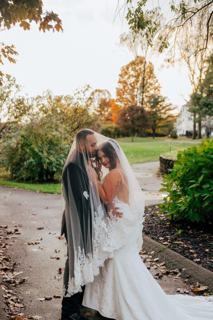 Groom kissing bride on the head under wedding veil outside during fall wedding at Black Swan Country Club in Georgetown, Massachusetts.