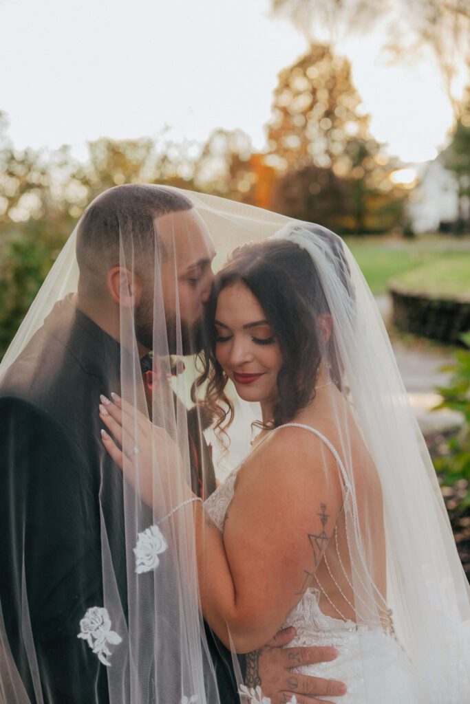 Groom kissing bride underneath bridal veil as she smiles outside at Black Swan Country Club in Georgetown, Massachusetts.