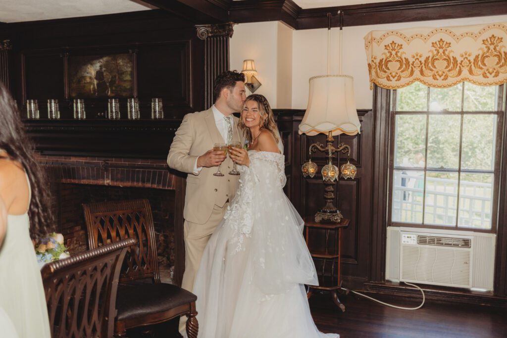 Groom is kissing bride as they are holding wine glasses in a charming room at Peirce Farm Estate in Topsfield, Massachusetts.
