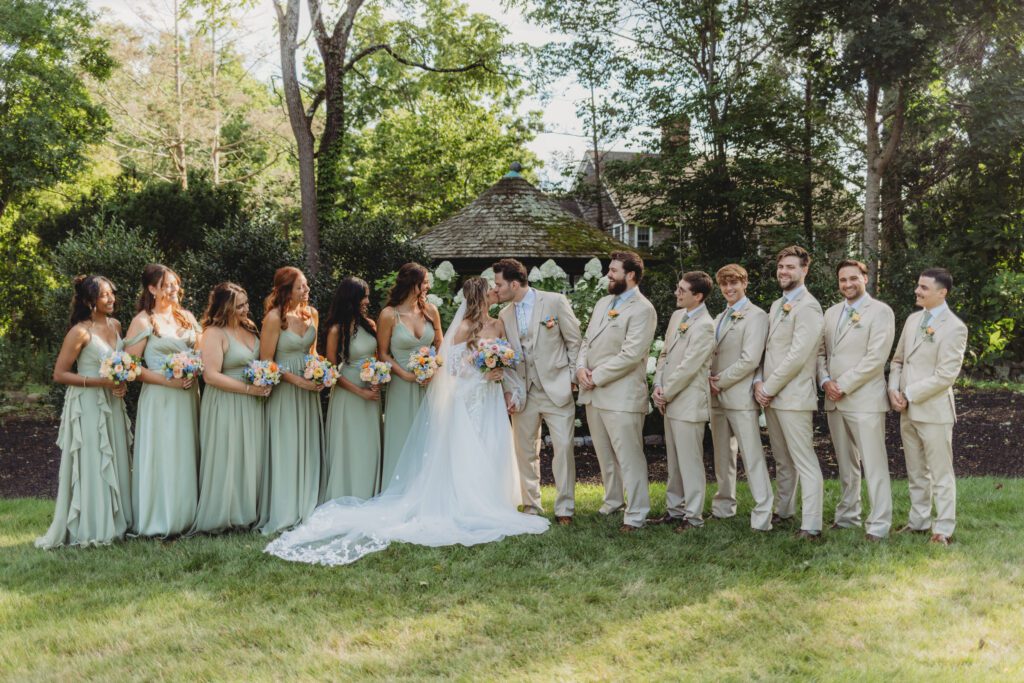 Bridal party standing together as bride and groom kiss outside at Peirce Farm Estate in Topsfield, Massachusetts.