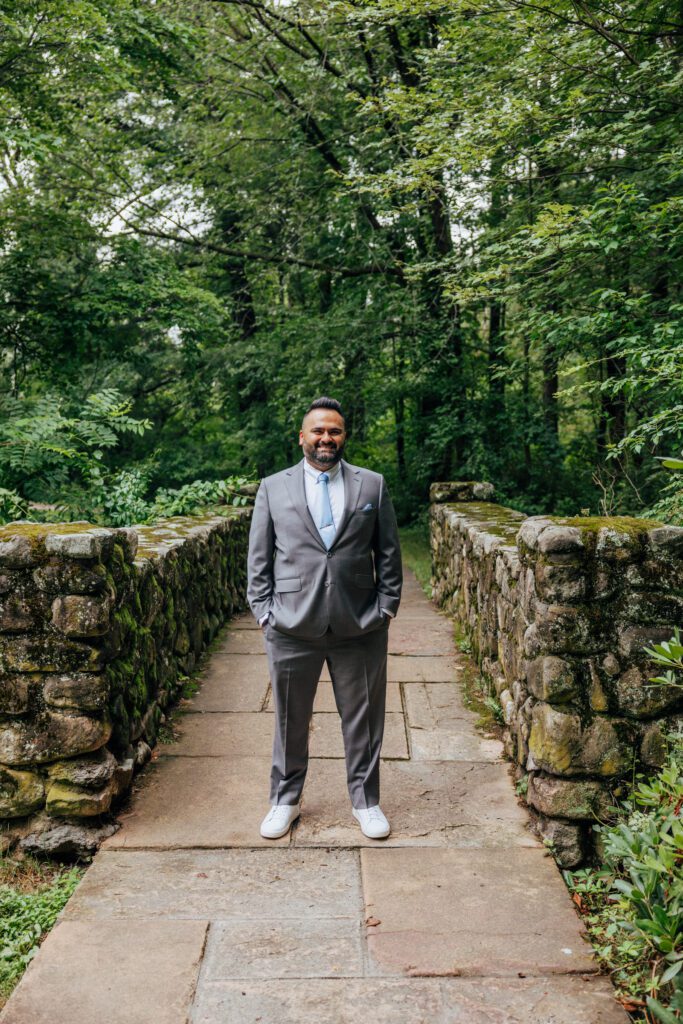 A groom smiling and standing on a stone bridge in the middle of the woods at Bird Park in Walpole, Massachusetts.