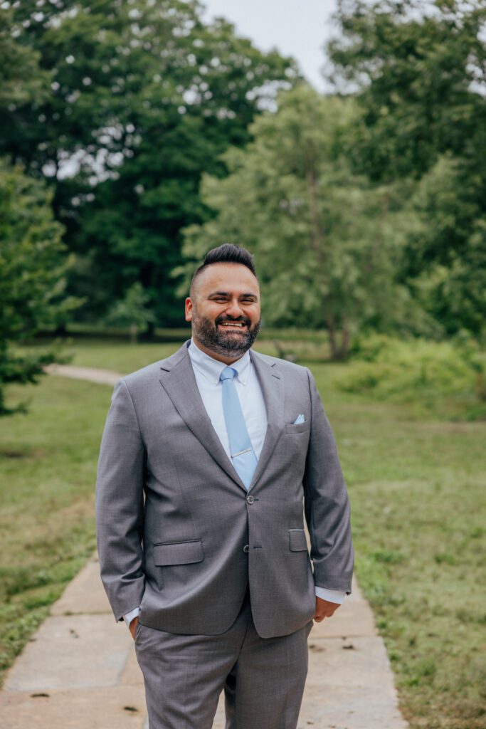 Groom smiling in the middle of a pathway surrounded by lush greenery in the summer at Bird Park in Walpole, Massachusetts.