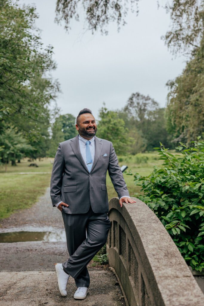 Groom laughing as he stands on a bridge in the summer at Bird Park in Walpole, Massachusetts.