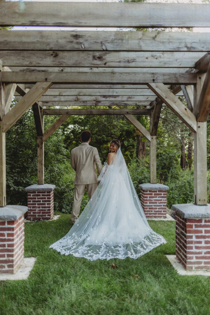 A smiling bride looks back over her shoulder at the groom walking away under an outdoor archway at Peirce Farm Estate in Topsfield, Massachusetts.