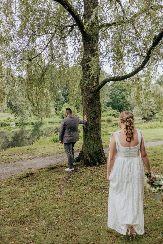 A bride is walking towards groom as he leans against a tree to have a first look outside at Bird Park in Walpole, Massachusetts.