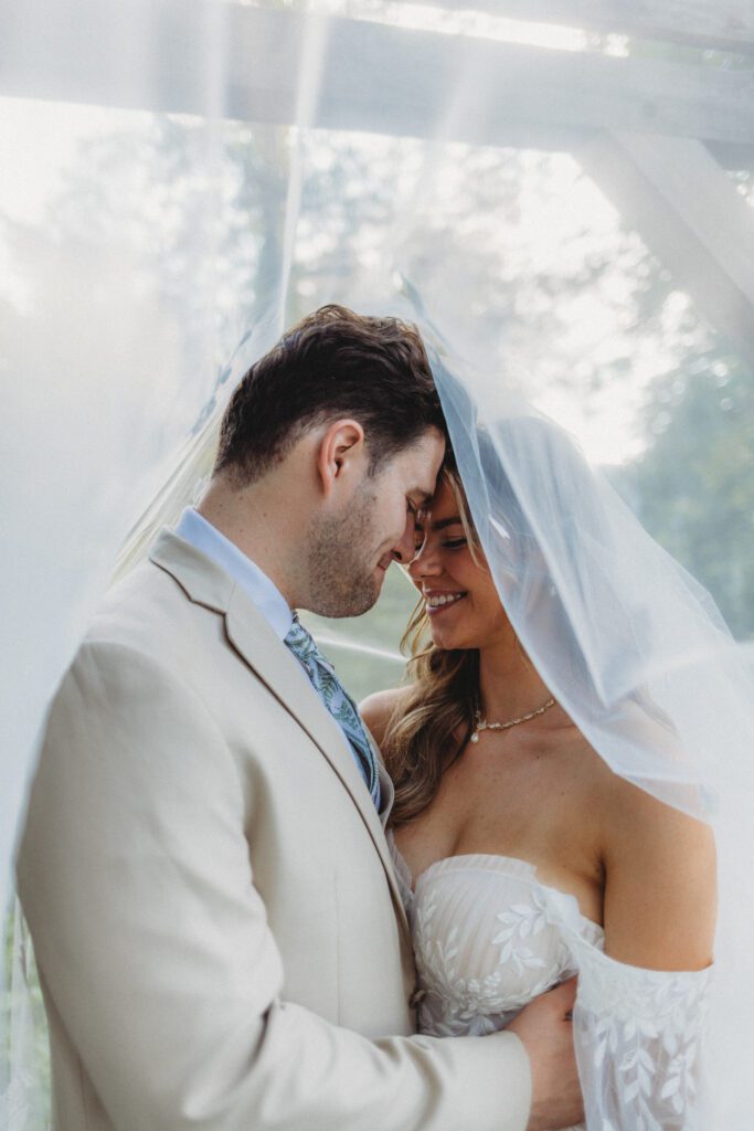 A bride an groom romantically smiling under a wedding veil at Peirce Farm Estate in Topsfield, Massachusetts.