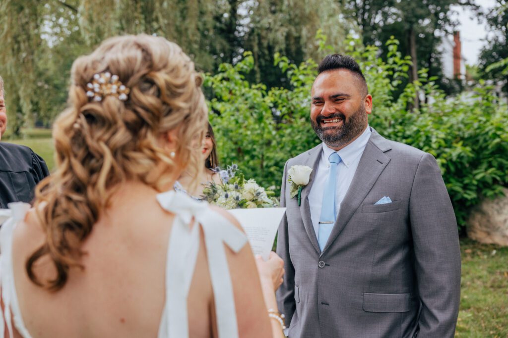 Groom smiling at bride as she reads her vows during romantic summer elopement at Bird Park in Walpole, Massachusetts.