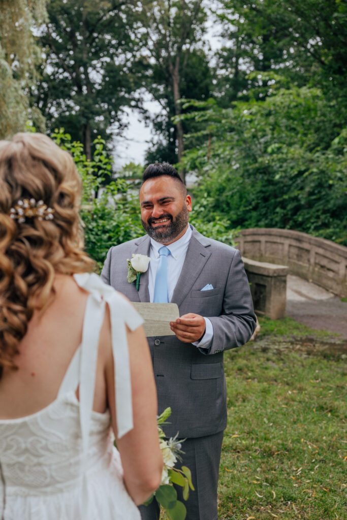 Groom smiling at bride reading vows during summer elopement ceremony at Bird Park in Walpole, Massachusetts.