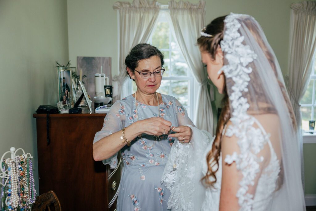 Mother of the bride fixing necklace for bride as bride is getting ready for Massachusetts wedding.
