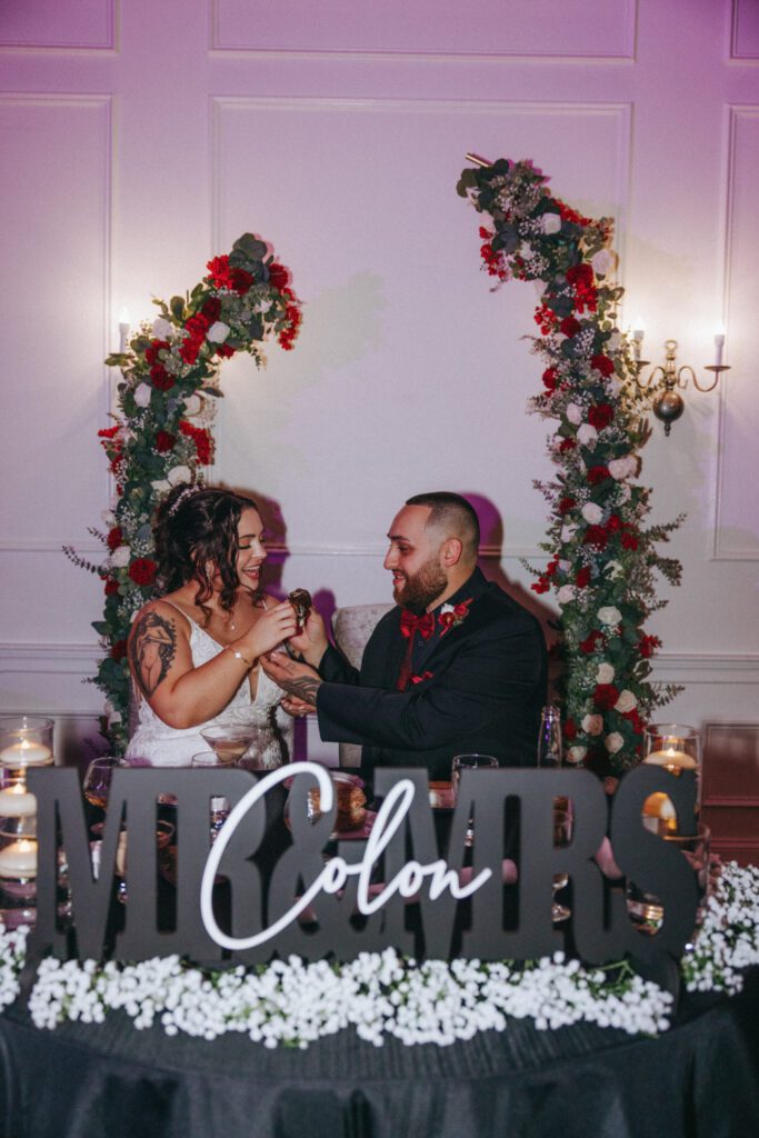 Bride and groom eating chocolate covered strawberries under flower arch at Black Swan Country Club in Georgetown, Massachusetts.